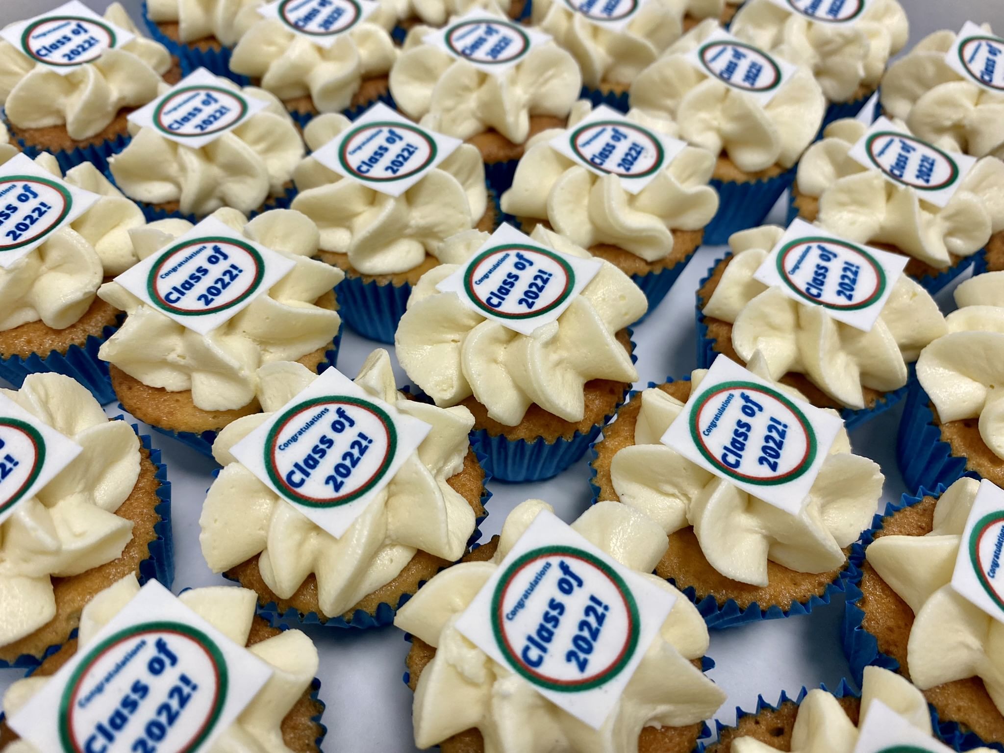 A large assortment of Mini Vanilla Cupcakes with cream-colored frosting, each topped with a circular edible decoration that reads 'Class of 2022!' in blue text. Perfect for corporate events, the frosting is piped in a floral pattern, and the cupcakes are in blue paper liners.